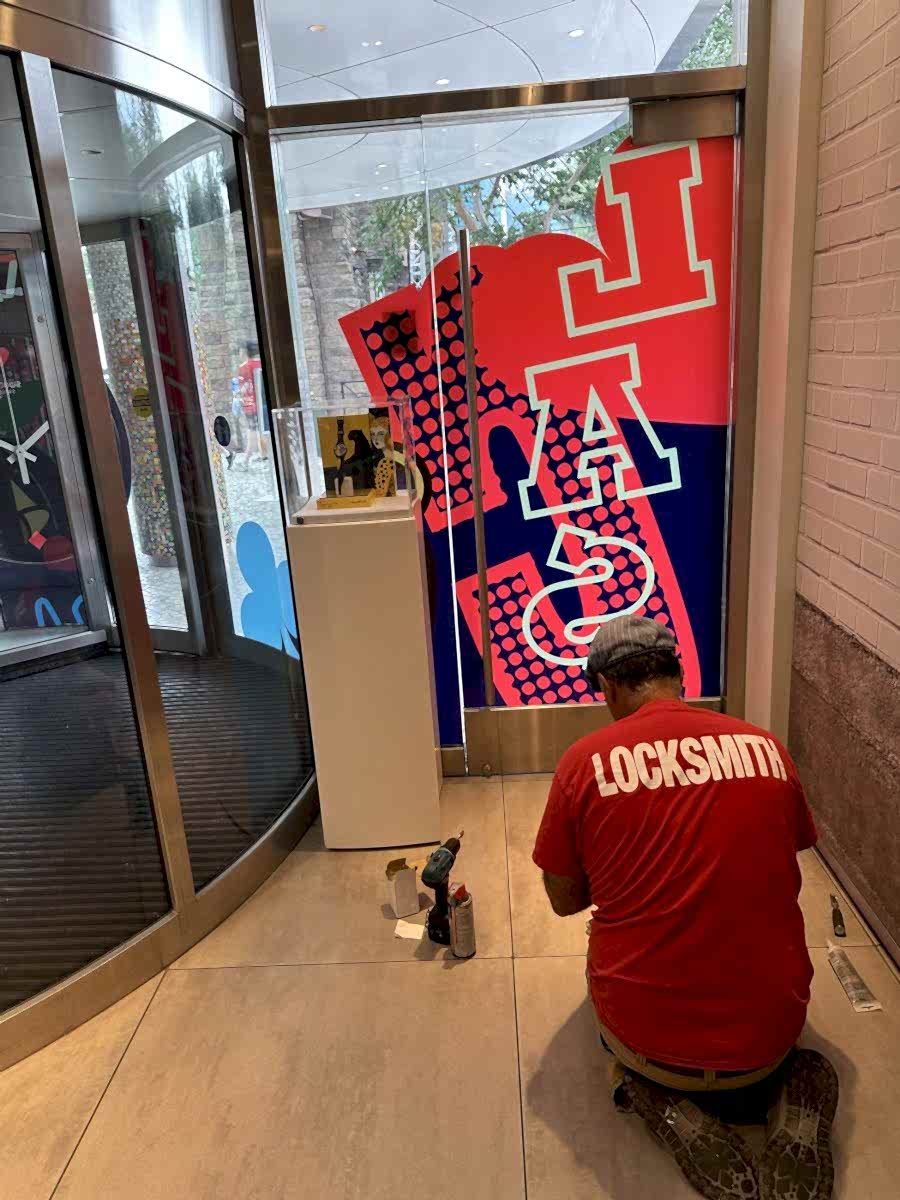 Locksmith in a red shirt kneeling at a glass storefront entrance with tools on the floor.